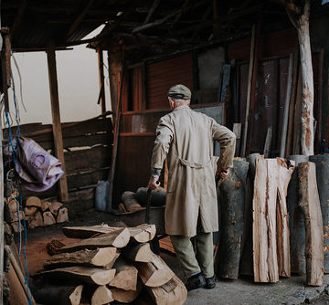 Old man in trench coat carrying firewood with a wheelbarrow.